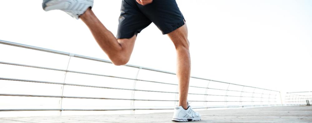 Henkilön jalat juoksemassa laiturilla, mustat shortsit ja valkoiset lenkkitossut, kirkas taivas taustalla.