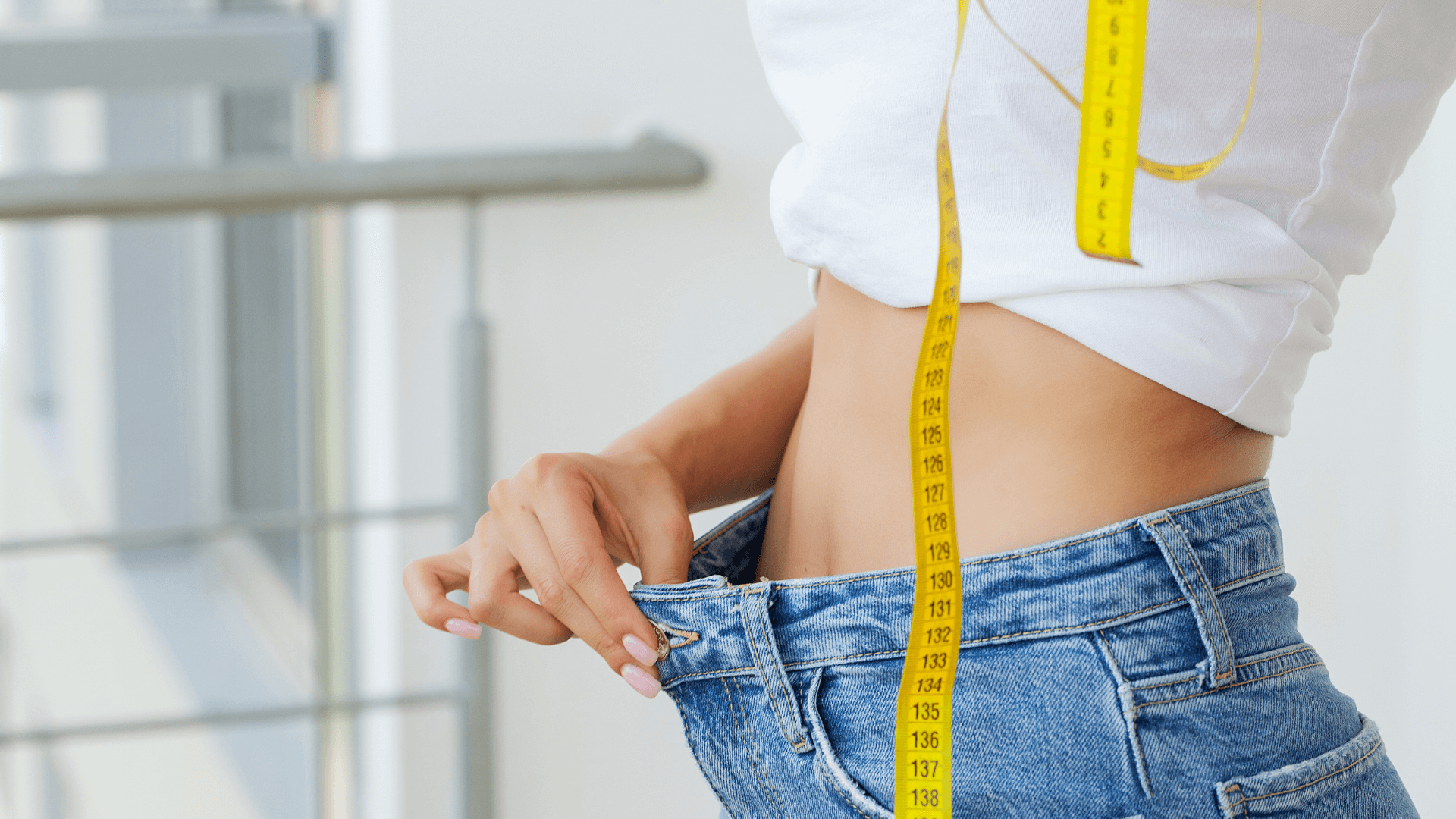 Nainen mittaamassa vyötäröään. Farkut ovat löysät laihdutuksen myötä. Kädessä keltainen mittanauha.